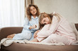 © Cookie Studio - Best friend cheer up heartbroken blonde. Portrait of two attractive women sitting on sofa in nightwear. Curly-haired girl drinking coffee, patting sister on head while she is upset or feeling cramps