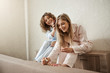 © Cookie Studio - Spending weekend with sister is better than alone. Charming happy curly-haired woman in pyjamas combing her friend hair while she is painting fingernails on feet, laughing and talking about life
