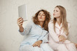 © Cookie Studio - Say mwah to camera. Two beautiful girlfriend sitting at home in nightwear having fun while taking selfie with digital tablet, folding lips as if sending air kiss, expressing friendliness and happiness