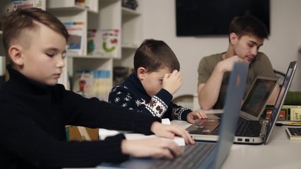 Two little boys typing something on laptop keyboards while their male programmer teacher is sitting near.