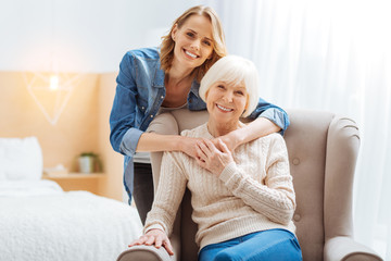 Close people. Positive emotional elderly woman sitting in a soft comfortable armchair while her kind attentive loving granddaughter standing behind her back and softly hugging her