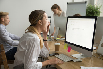 Tired businesswoman yawning working on computer sitting at desk with colleagues, sleepy employee gaping suffering from lack of sleep, feeling bored or chronic fatigue in office concept, side view