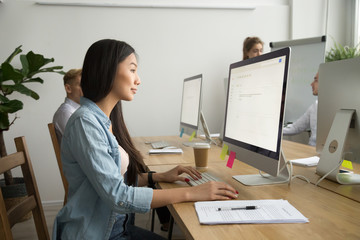 Young asian businesswoman working online on pc desktop computer in modern multiracial office, chinese intern employee using applications typing business email in corporate coworking, side view