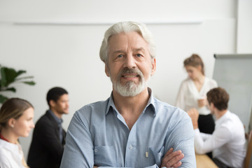 Confident senior businessman leader looking at camera with team at background, smiling aged company boss posing in office with arms crossed, older mentor or executive professional head shot portrait