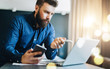 © foxyburrow - Young bearded businessman is sitting in front of computer, holding smartphone and showing pen on laptop screen. Freelancer, entrepreneur works at home. Distance work, online education, marketing.
