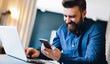 © foxyburrow - Young smiling bearded businessman sitting at table in front of computer, using smartphone. Freelancer, entrepreneur works at home. Online marketing, education for adults, distance work. Social network