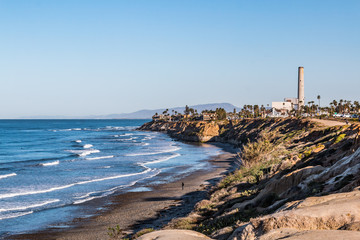 Man walks near cliffs on South Carlsbad State Beach in San Diego, California with the power plant landmark tower in the background. 