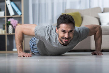 Portrait of smiling strong guy in living room doing push ups on floor