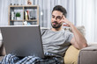 Portrait of pleased bearded man relaxing on sofa with notebook. Bookshelf on background