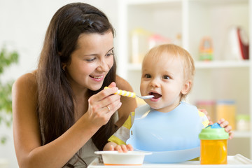 Poster - Mommy giving healthy food to her baby son on high chair in kitchen.