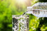 Fototapeta Łazienka - Drinking water pouring from bottle into glass on blurred fresh green nature background, healthy drink concept