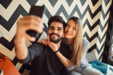 Wall Mural - Happy young couple taking selfie with smart phone at cafe in mall. Copy space for your text. Shallow depth of field. Very useful photo for processing with one click on edit image.