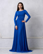 Young woman with long brunette hair dressed in elegant evening blue maxi dress with long sleeves posing in studio. Gorgeous female model standing against white wall decorated with classic moldings.