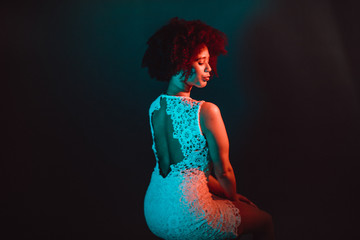 Wall Mural - Beautiful black woman with curly hair showing her naked back, wearing a white dress in a studio in front of a dark background