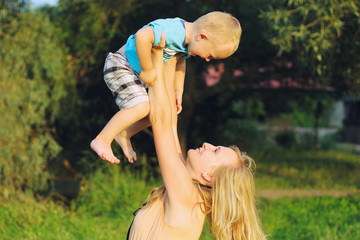 Young blonde mother in playing with her child in the green lawn. Happy fun summer photo. Smiling laughing boy with his mom. Sunny day outdoors in nature.