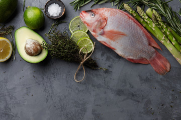 Poster - Raw red tilapia fish cooking with herbs, spices, green asparagus, avocado, lemon and lime on gray stone background, top view