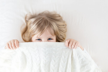 Little girl peeking out blanket on bed. Cute kid smiling and hiding under knitted cover. Palyful and mischievous eyes. Hide-and-seek. Children having fun playing active games. Happy childhood concept