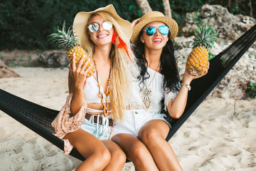 Two cute beautiful girls in boho style dressed shirts shorts light cape bracelets necklaces, in the hair decorative colored feathers, straw hats, with pineapple in their hands sit in a hammock