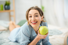 Portrait Of A Young And Cute Woman Lying With Green Apple In The Cozy And Bright Bedroom At Home