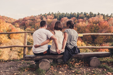 Three young beautiful people sit on a wooden bench and enjoy the autumn landscape, the concept of a love triangle and betrayal