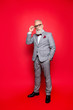 © deagreez - Portrait of cool, virile, harsh, perfect old man in checkered grey suit and eyewear, standing, isolated on red background, looking at camera, holding hand on eyelet of glasses on his face
