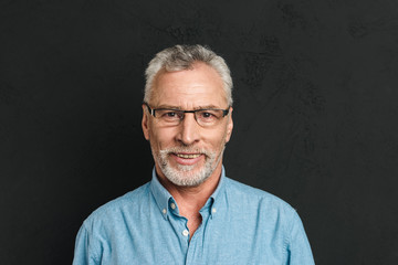 Horizontal photo of mature unshaved man 60s with grey hair wearing eyeglasses smiling and looking on camera with candid gaze, isolated over black background