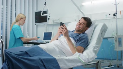 Wall Mural - Recovering Patient Uses Smartphone and Smiles while Lying on a Bed in the Hospital. Friendly Nurse Comes in to Check on Him.