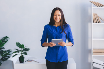 young woman holding digital tablet, smiling and looking at camera while standing at modern office.