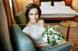 Elegant charming young brunette bride in the hotel room  is sitting on sofa in a wedding dress with a big bouquet of flowers, daydreaming, in the interior Waiting for groom. detailed retouch