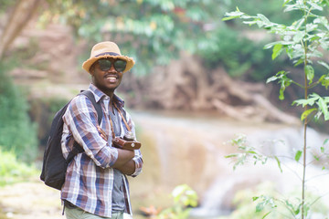 Wall Mural - African Man Traveler with backpack standing and holding camera with waterfall background.