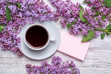 White Cup With Coffee Or Tea, Note With Wishes, A Bouquet Of Lilacs On A Wooden Background. Violet Spring Flowers. Morning Composition.