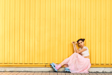 Young Girl In Pink Skirt And Roller Skates Sits On Sidewalk Against Yellow Wall Background And Hands Makes Heart. Concept Life Style. Copy Space