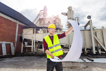 Wall Mural - Young engineer or worker wearing yellow safety helmet and vest holding construction plan