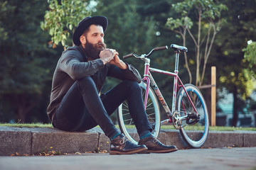 Wall Mural - A handsome hipster traveler with a stylish beard and tattoo on his arms dressed in casual clothes and hat with a bag, sitting on the sidewalk, resting after a bike ride.