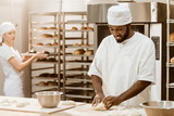 Fototapeta Do przedpokoju - handsome african american baker kneading dough while his female colleague working on background at baking manufacture