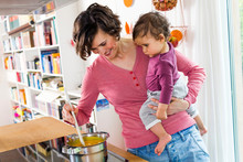Mother Holding Baby Girl In Kitchen, Whilst Stirring Pot On Stove