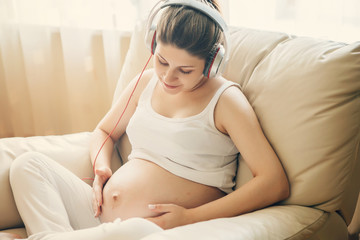 Portrait of pregnant woman sitting on sofa at home and listening music in headphones.