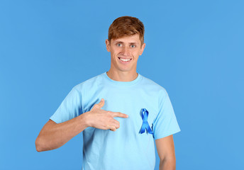 Wall Mural - Young man pointing at blue ribbon on his t-shirt against color background. Prostate cancer awareness concept