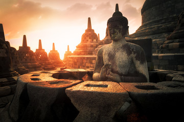 Wall Mural - Amazing sunrise view of meditating Buddha statue and stone stupas against shining sun on background. Ancient Borobudur Buddhist temple. Great religious architecture. Magelang, Central Java, Indonesia