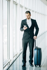 Happy young businessman walking and looking at mobile phone at airport. Handsome business executive texting on smartphone while walking in airport terminal.