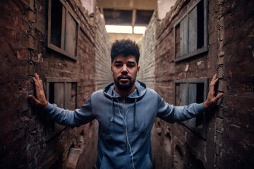 Portrait of focused motivated afro-american young attractive athletic man with earphones standing inside of the abandoned place in the middle of two walls and looking at the camera.