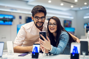 Wall Mural - Close up focus view of a stylish charming embracing young student love couple taking a selfie with new mobile in a tech store.