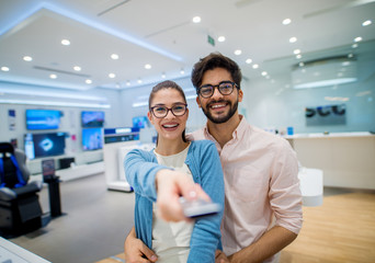 Close up portrait view of cute excited charming young student girl with eyeglasses holding tv remote control while her boyfriend hugging her in a tech store.