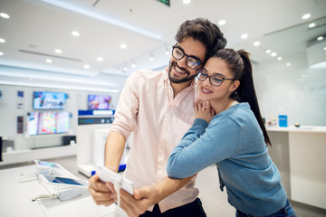 Wall Mural - Portrait view of young cute love couple standing hugged in front of the desk with tablets and testing selfie operation in the tech store.