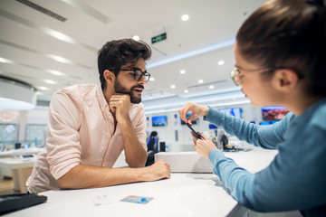 Close up portrait view of curious excited happy smiling young student bearded man looking at mobile while female seller giving instruction for sim place on the phone.