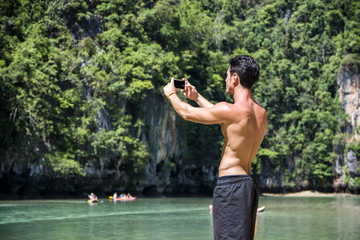 Wall Mural - Half body shot of a handsome young man using cell phone, standing on a beach in Phuket Island, Thailand, shirtless wearing boxer shorts, showing muscular fit body