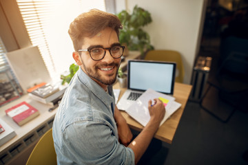 Wall Mural - Portrait of happy motivated smiling hipster student learning for a test or an exam at high school library desk while sitting turn backwards and looking at the camera.