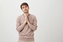 Portrait Of Cute Adult Man With Bristly Holding Hands On Chest In Pray Gesture, Begging For Something With Adorable Look, Standing Over Gray Background. Son Asks Mom To Come Home Late