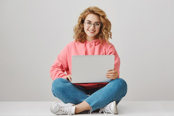 Wall Mural - Help me with homework. Portrait of happy interesting girl with curly hair, working on project, sitting on floor with crossed legs, holding laptop and smiling broadly, freelancing over gray background