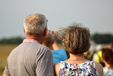 Fototapeta  - Para osób w dojrzałym wieku, kobieta i mężczyzna, stoją tyłem, w plenerze, patrzą na coś współnie, prezd nimi nieostra postać męzczyzny, wiatr rozwiewa kobiecie krótkie blond włosy